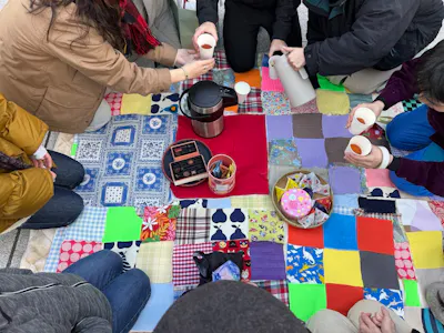 ワークショップのイメージ写真。7，8人の人々が円になって飲み物を乾杯してピクニックを楽しんでいる